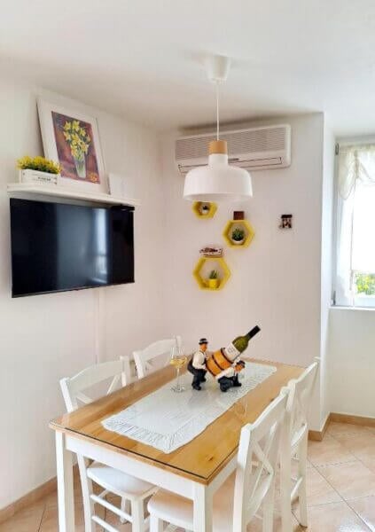 A small dining area at Megi Apartman on Brac island with a wooden table set for one, white chairs, a wine bottle holder centerpiece, wall-mounted TV, hexagonal shelves, and a window letting in natural light.