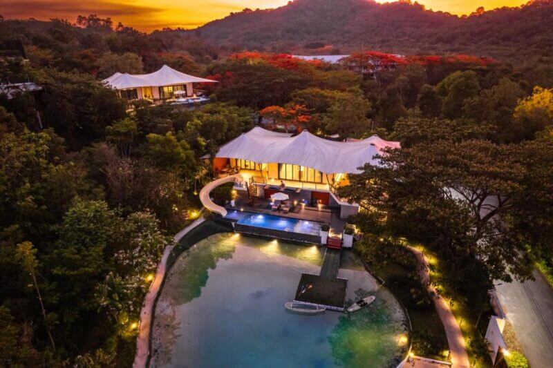Aerial view of a luxury villa with a pool at Te Mata Glamping, surrounded by dense trees and Khao Yai hills at sunset, with warm lighting illuminating the property for an unforgettable glamping experience.