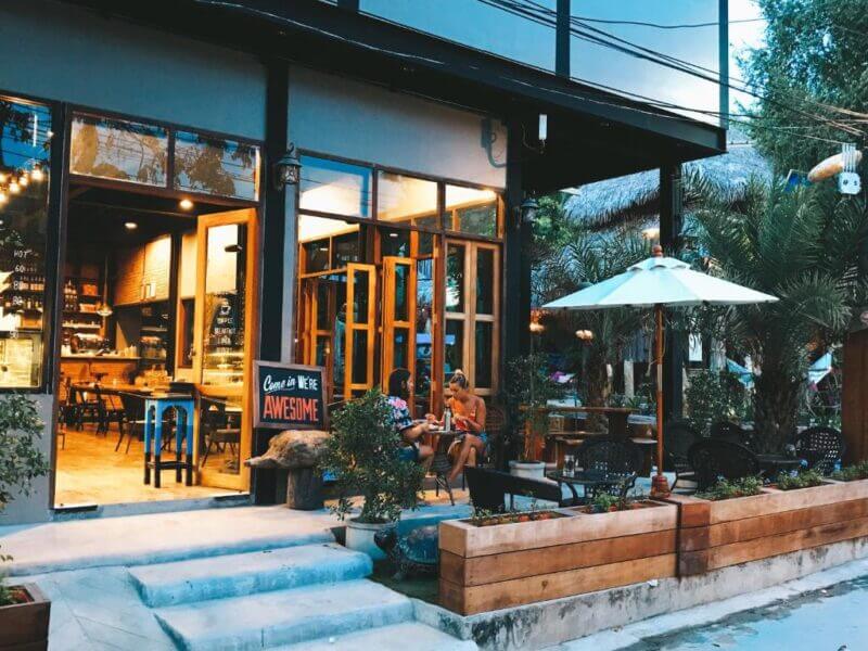 Two people sit at an outdoor table in front of Bloom Hostel’s modern café in Koh Lipe, with large windows, lush plants, and a cozy umbrella while the warmly lit interior invites guests inside.