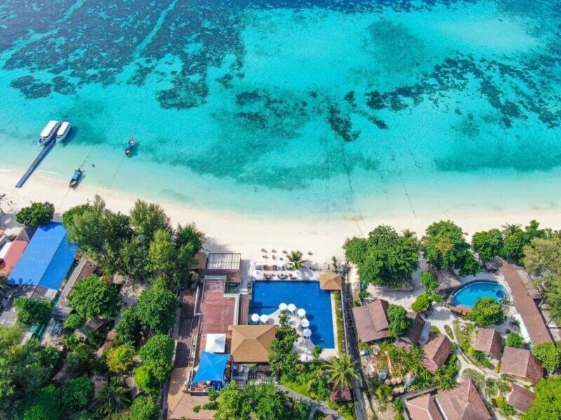 Aerial view of AKIRA Koh Lipe showcases a tropical beach with clear blue water, anchored boats, a swimming pool, and several buildings surrounded by trees along the stunning Koh Lipe shore.
