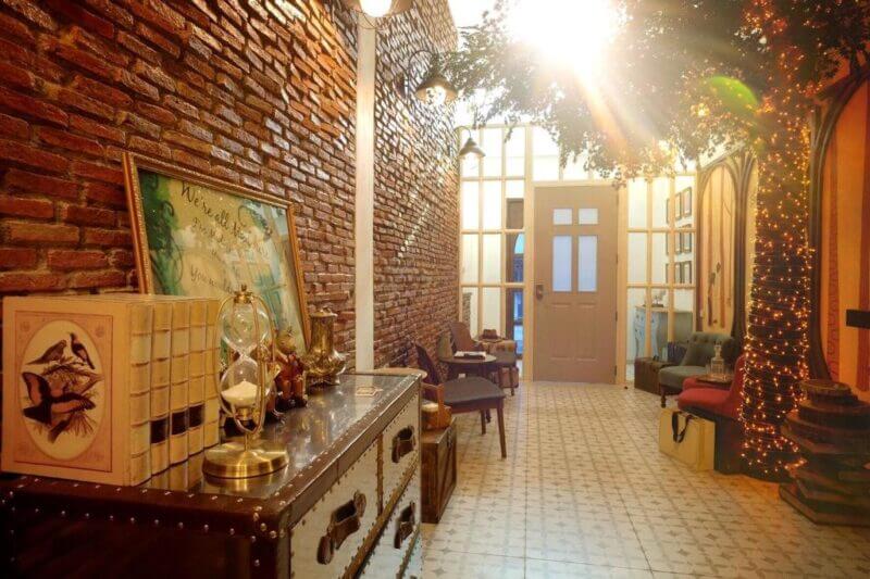 A warmly lit hallway with exposed brick walls, vintage furniture, a large potted tree adorned with string lights, and sunlight streaming through glass doors at the far end—inviting you to discover the charm of Hide and Seek Bangkok.