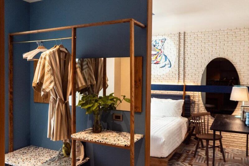 A stylish room at Tints of Blue Bangkok featuring a blue accent wall, open wooden closet with striped robes, a mirror, and a bed with crisp white linens in the background.