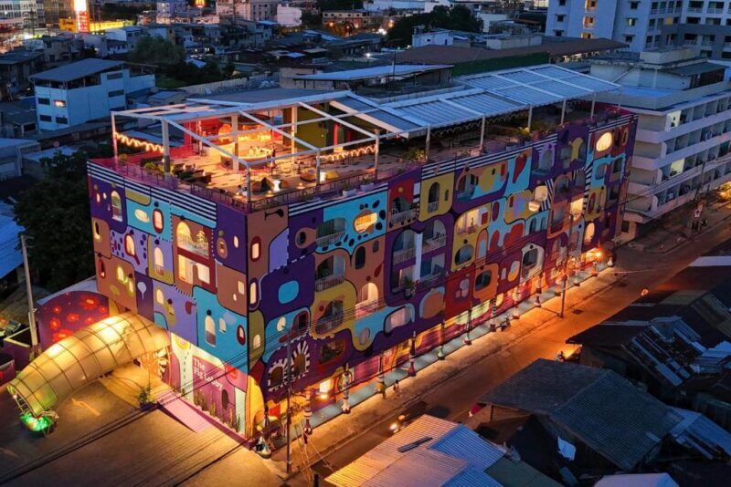 A multistory building with a colorful, abstract mural facade, rooftop seating area, and lit street in an urban setting at dusk, reminiscent of the vibrant energy found at Fig Lobby Bangkok.