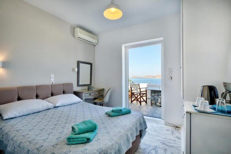 A neatly made bed with teal towels, a desk with a mirror, and a door open to a balcony with chairs overlooking the sea and distant hills—enjoy relaxing accommodation at Irene Rooms in Naoussa.