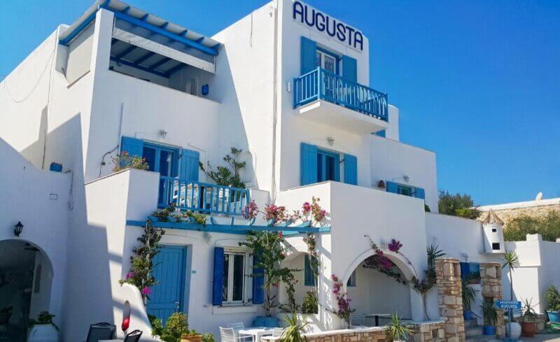 White Mediterranean-style building with blue shutters and balconies, labeled Augusta Studios, nestled in Naoussa and surrounded by flowering plants under a clear blue sky.