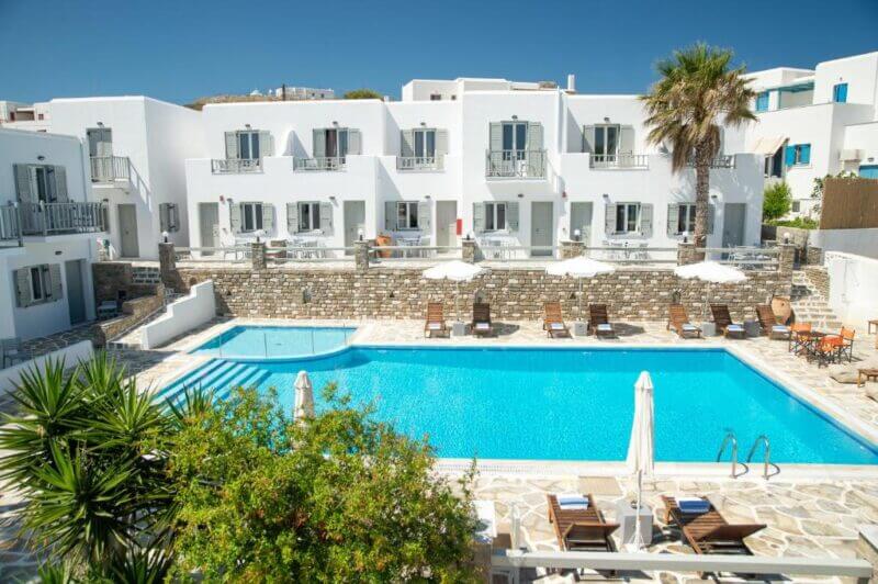 A large outdoor swimming pool is surrounded by sun loungers and umbrellas in front of the white, two-story Hotel Arkoulis with balconies and green shutters, offering a relaxing retreat in Naoussa under a clear blue sky.