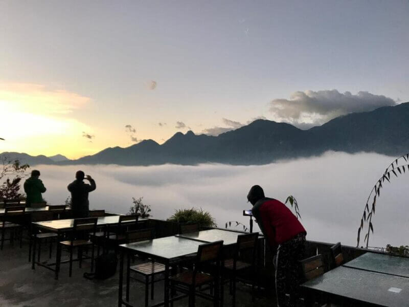 Three people stand on a terrace at their Sapa homestay with empty tables, taking photos of misty Fansipan mountains at sunrise or sunset.