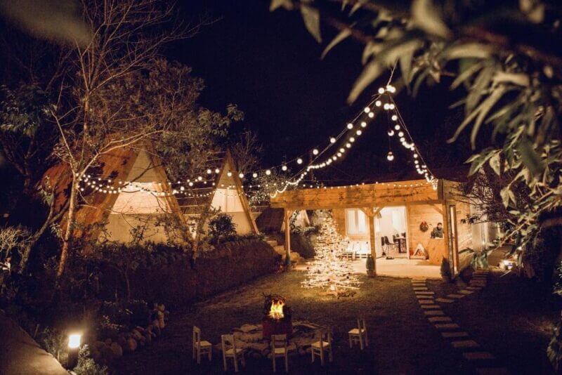 Outdoor nighttime scene at TEEPEE Homestay in Dalat, with string lights overhead, a fire pit surrounded by chairs, a gazebo, and several triangular cabins nestled in a charming garden setting.