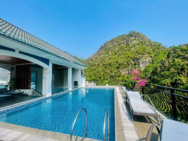 Enjoy the outdoor swimming pool at Cat Ba Villa with lounge chairs, a covered seating area, and stunning mountain views in the background under a clear blue sky in Cat Ba.