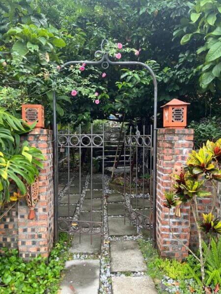 A brick and metal garden gate with arch and pink flowers leads to a stone path surrounded by lush green plants and trees, reminiscent of a tranquil Blue Lagoon retreat.