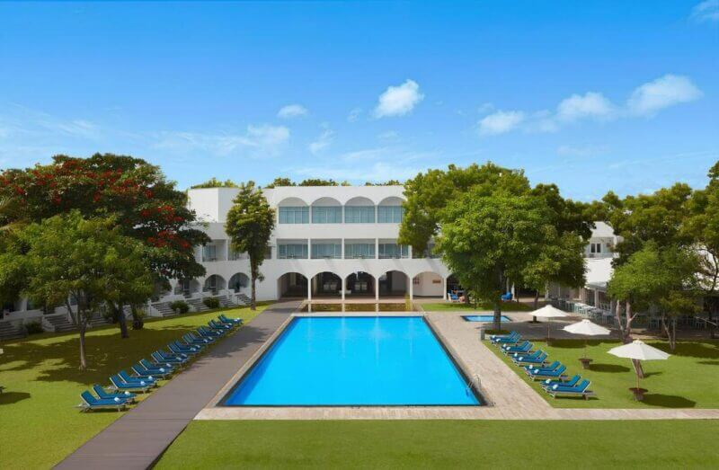 A rectangular outdoor swimming pool at Trinco Blu is surrounded by lounge chairs and umbrellas, with a white, two-story building and lush green trees in Trincomalee set against a clear blue sky.