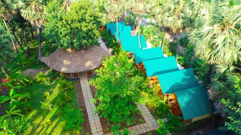 Aerial view of a lush garden at Sasvi Cabana Trincomalee, featuring a thatched pavilion and teal-roofed buildings, surrounded by palm trees and green pathways—one of the most serene Trincomalee hotels.