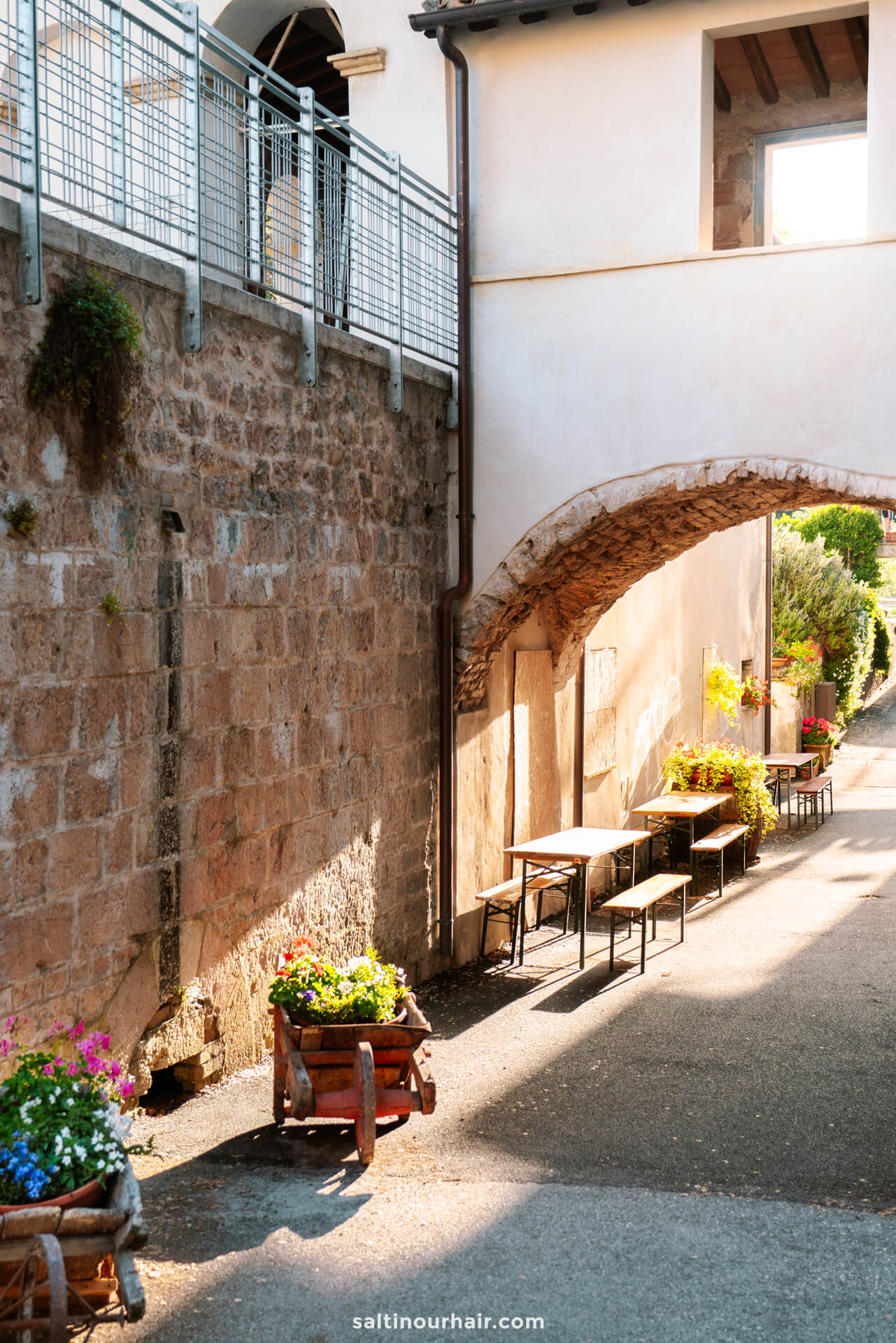 A narrow outdoor walkway in Rasiglia, Umbria, Italy, with tables, benches, potted flowers, and an arched stone passageway beside a sunlit stone wall.