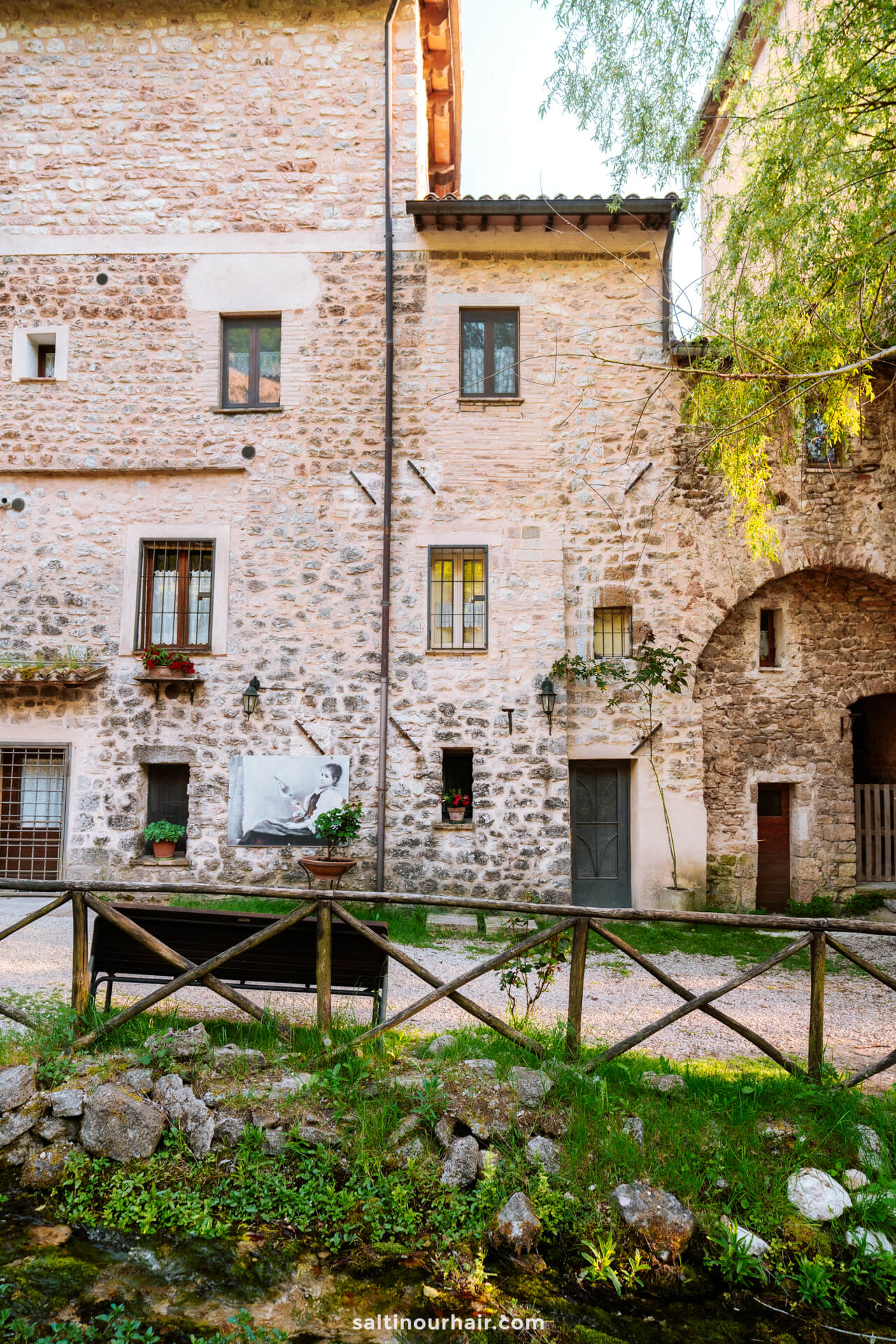 Stone facade of an old building in Rasiglia, Umbria, Italy, with various windows and doors, potted plants, a wooden fence, and a bench in front, surrounded by lush greenery.
