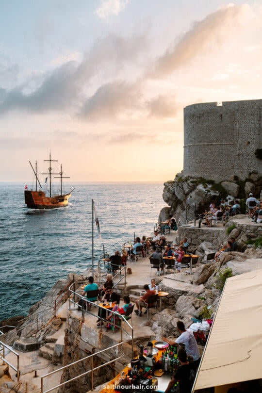 People sit at outdoor tables on rocky terraces overlooking the sea at sunset in Dubrovnik, with a large stone wall and a tall ship sailing nearby&mdash;an unforgettable scene from a road trip Croatia travelers will always cherish.