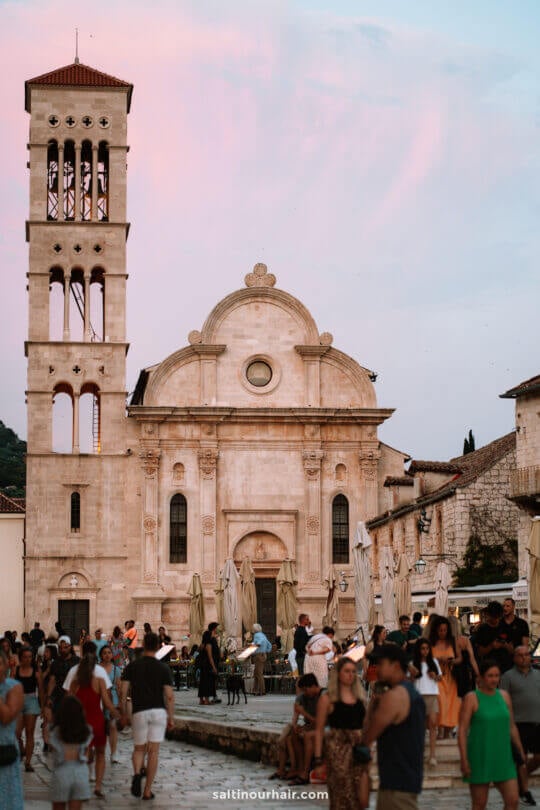 Crowds of people gather in front of a historic stone church in Hvar with a tall bell tower at sunset, enjoying outdoor seating and umbrellas in the square&mdash;a picturesque stop on any road trip Croatia adventure.