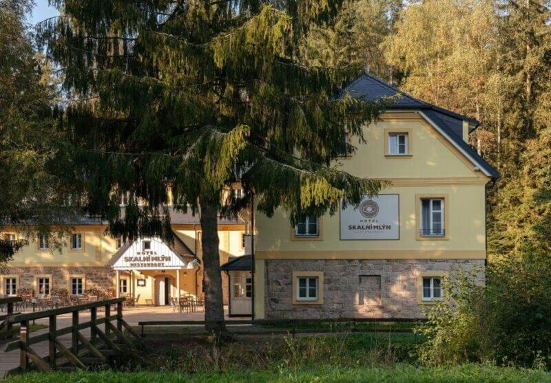The Skalní Mlýn hotel building stands with a large tree in the foreground, welcoming visitors on their way to explore the breathtaking Adršpach rocks.