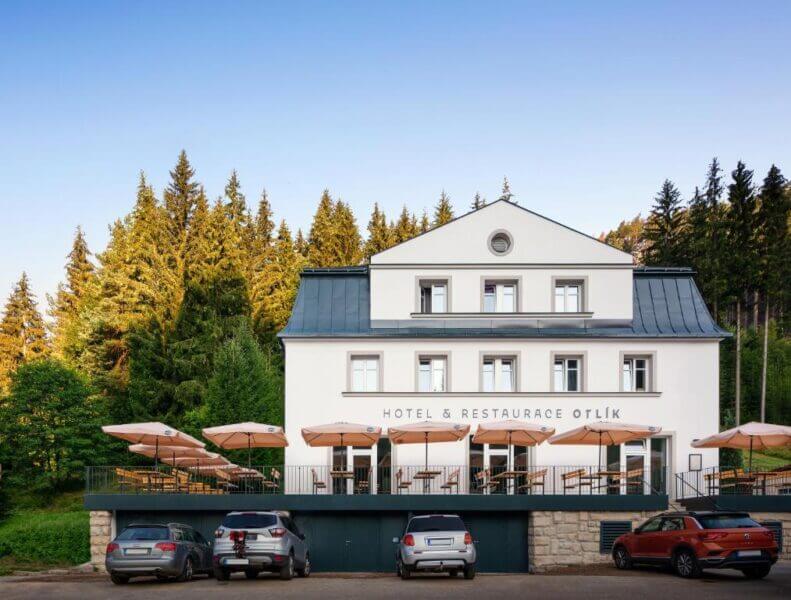 The Adrspach Hotel is a white two-story building with a restaurant, outdoor terrace seating, parked cars in front, and tall trees in the background under a clear blue sky.