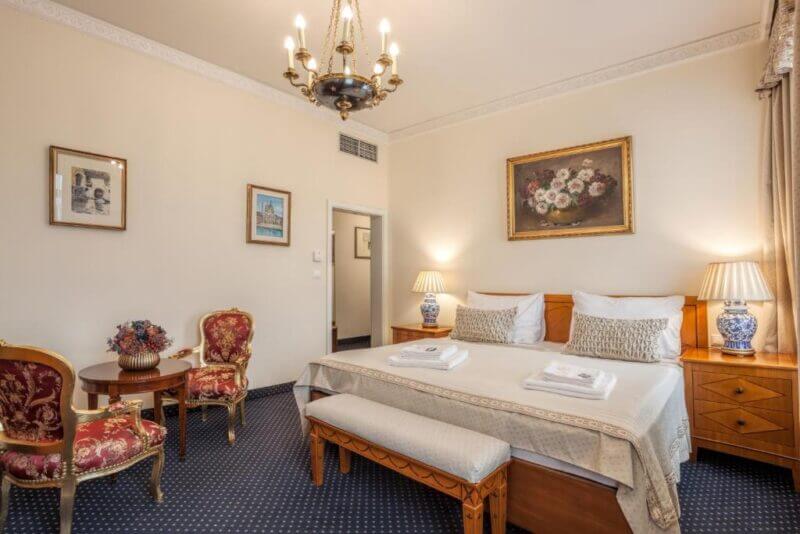 A Brno Hotel room featuring a double bed, two bedside tables with lamps, a cozy seating area with chairs and table, framed artwork on the walls, and a chandelier overhead—an inviting Brno accommodation at Hotel Europa.