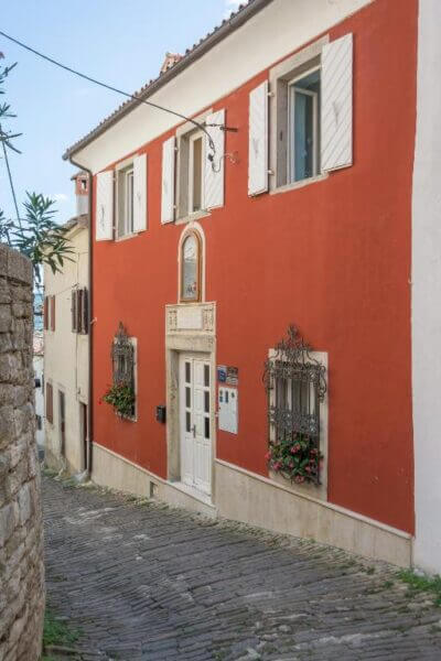 A narrow stone street passes by Villa Marija Guesthouse, a two-story building in Motovun with a red facade, white shutters, arched doorway, and window flower boxes.