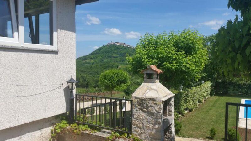 A stone gate and fence beside a white guest house, with green trees and the hilltop village of Motovun visible in the distance under a blue sky.
