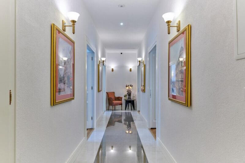 A well-lit hallway at Casa Dei Fiori in Pula features framed artwork on white walls, elegant sconces, and a polished floor leading to a table with chairs and stylish decor at the end.