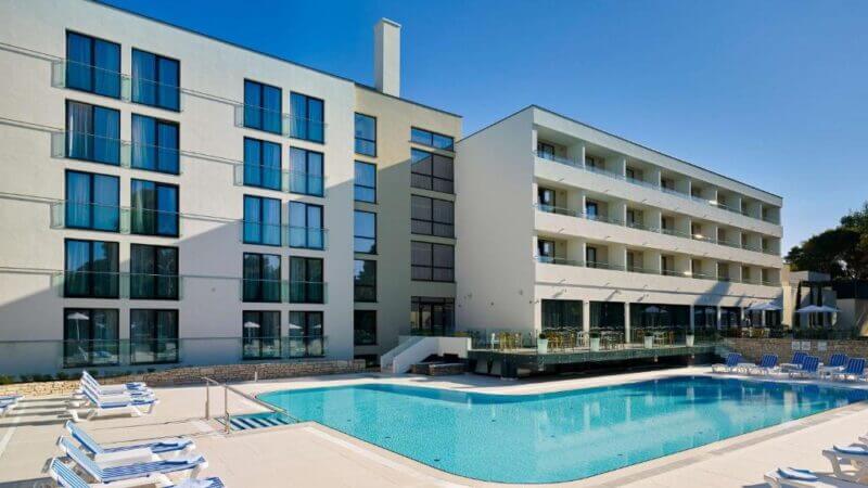 Modern hotel building with multiple floors and balconies, situated beside an outdoor swimming pool surrounded by lounge chairs under a clear blue sky at Park Plaza Arena hotel in Pula.