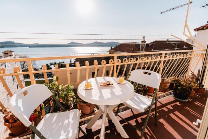 A small balcony at Arya Apartment features a round table, two chairs, potted plants, and drinks, overlooking Sibenik’s rooftops and a shimmering body of water with hills in the distance.