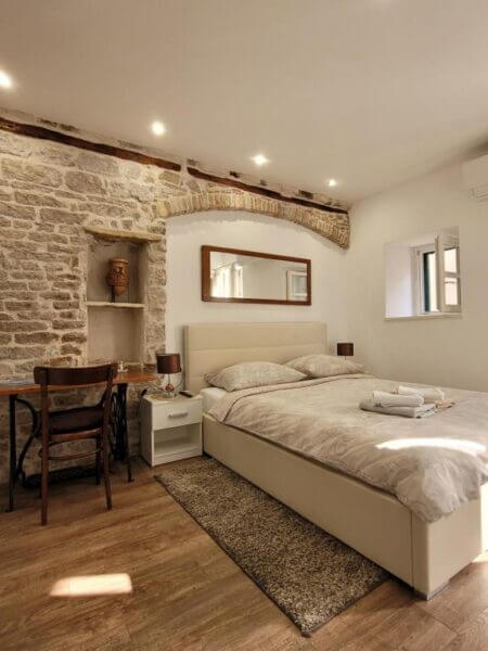 A luxury room in Sibenik featuring a double bed, two nightstands, a desk with chair, stone accent wall, window, and a mirror above the bed. Fresh towels are elegantly folded on the bed.