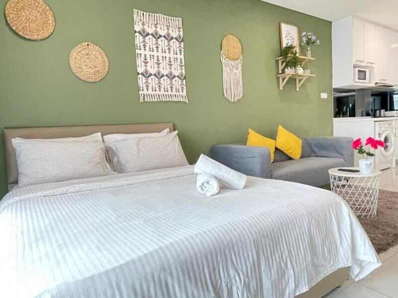 A neatly made bed with white linens sits beside a gray sofa with yellow pillows in a green-walled room at Mercu Summer Suite, Kuala Lumpur, featuring wall decor and a small white table topped with pink flowers.