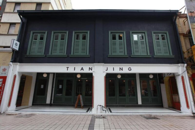 Two-story building in KL with dark upper walls, green shuttered windows, and a white lower facade labeled Tian Jing Hotel; glass doors on the ground floor open onto a tiled sidewalk.