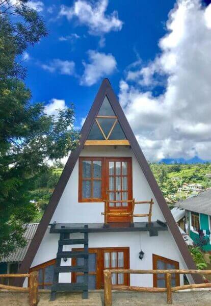 A-frame house with white walls and wooden trim, featuring a balcony and windows, set against a partly cloudy sky and green landscape—perfect for a peaceful escape at La Luna Cabins, Nuwara.