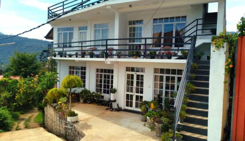 Two-story white house in Nuwara Eliya with large windows, balconies, black railings, and exterior stairs, surrounded by potted plants and lush greenery.