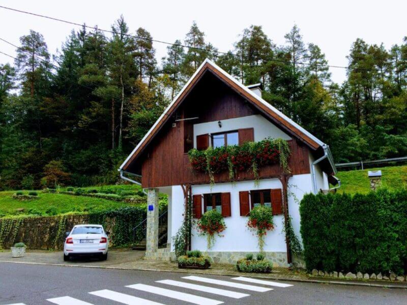 A white and brown two-story holiday house with flower boxes stands beside a road with a crosswalk in Rastoke; a white car is parked in the driveway, and forested trees provide a picturesque backdrop.