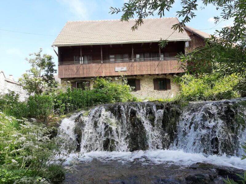 A rustic house with a wooden balcony and tiled roof stands behind a small, cascading waterfall surrounded by greenery in Rastoke, near Sobe Ivan at Rastoke 21.
