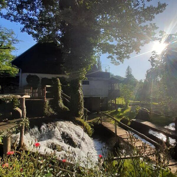 A rustic guesthouse with ivy-covered walls stands beside a small waterfall and wooden bridge in Rastoke, with sunlight shining through the trees in a lush garden.