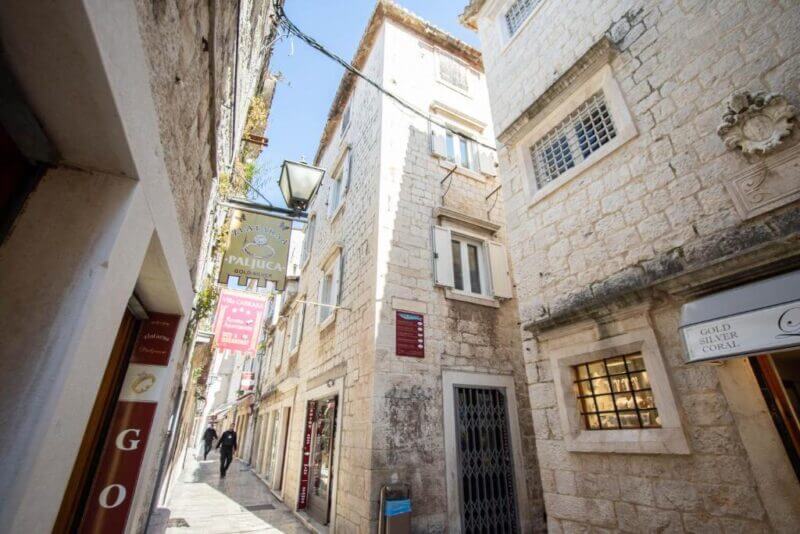 Narrow stone-paved street in Trogir lined with old stone buildings, shop signs, and a few pedestrians under a sunny sky—a charming scene near Stephane City Vibes Suites.