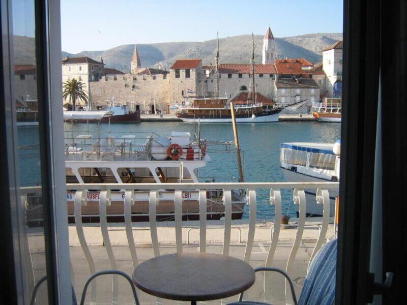 View from a window at Villa Tudor shows a small table and two chairs on a balcony overlooking boats docked in Trogir harbor, with historic stone buildings and hills in the background.
