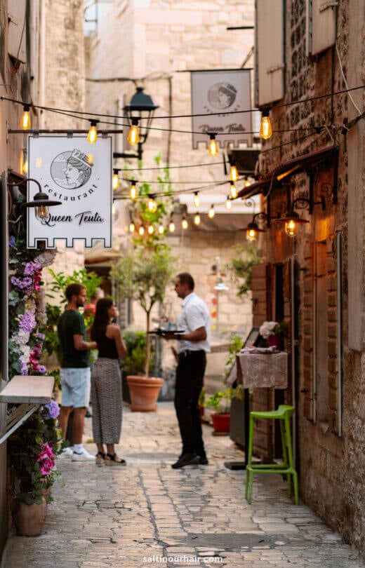 A waiter serves a man and woman standing in a narrow, cobblestone alley with hanging lights and Queen Teuta restaurant signs in Trogir&mdash;a charming stop on any 3-week Croatia itinerary.