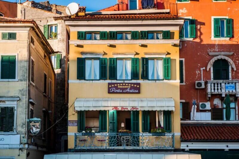 A yellow building with green shutters and a sign reading Residence Porta Rovinj stands between orange and beige buildings, featuring a balcony with potted plants and a striped awning.