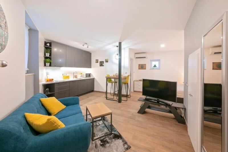 Modern Mariva Apartment in Rovinj featuring a blue sofa, yellow accents, compact kitchen, small dining area, and flat-screen TV on a stand in an open-plan layout with light wood flooring.