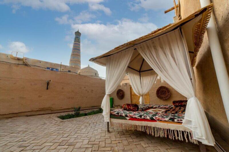 Outdoor seating area with cushions and curtains in a courtyard in Khiva, with the tall Islam Khodja minaret visible in the background under a partly cloudy sky.