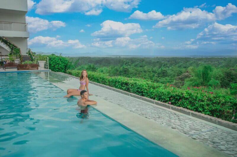 Two people relax at the edge of an outdoor infinity pool at Panja Resort, with lush greenery and the wide landscape of Puerto Princesa, Palawan visible under a blue, partly cloudy sky.