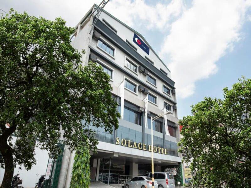 Exterior view of Solace Hotel, a Manila hotel with a sign above the entrance and several parked cars in front, surrounded by trees under a partly cloudy sky.