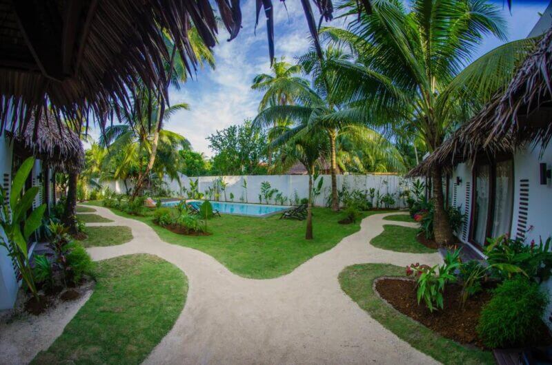 Tropical courtyard at Palaka Resort with a small swimming pool, palm trees, and sandy paths bordered by greenery between thatched-roof buildings under a partly cloudy Siargao sky.
