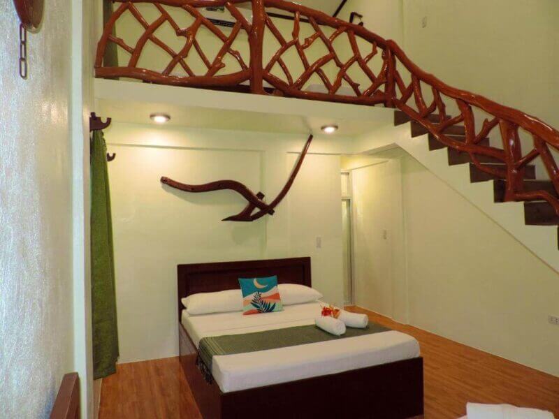 A modern Casa loft bedroom with a wooden staircase, double bed, green accents, and decorative wooden wall art above the bed, inspired by the relaxed vibe of Siquijor.