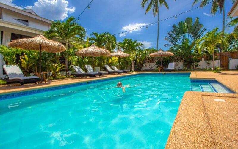 A person swims alone in a clear outdoor pool at Dolce Amore Resort, surrounded by lounge chairs, thatched umbrellas, palm trees, and tropical plants under a sunny Siquijor sky.