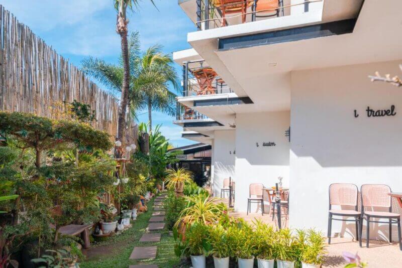 Outdoor walkway beside Bay Area Lodge, a modern white building with balconies, potted plants, outdoor seating, and a bamboo fence under a blue sky.