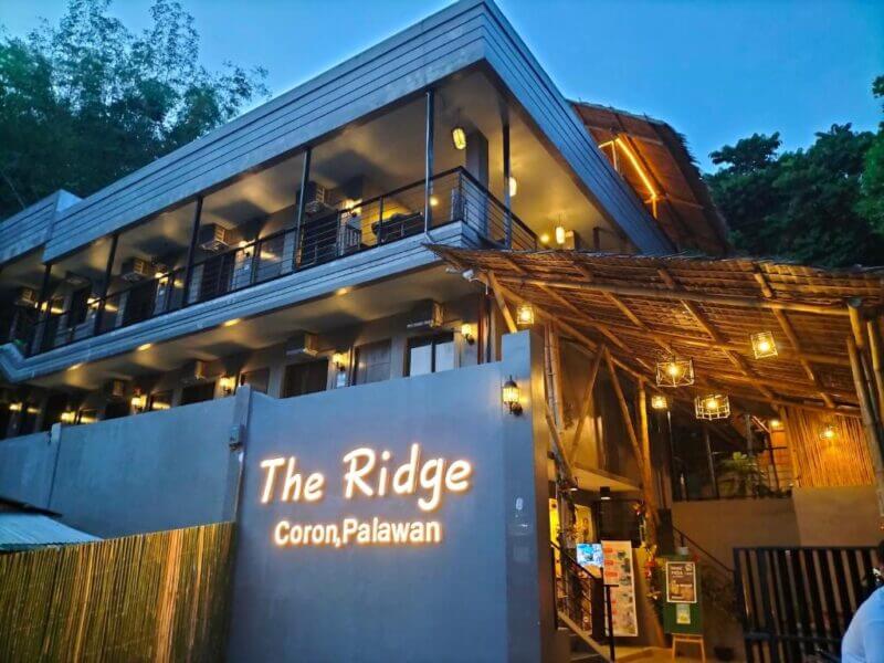 Modern two-story building with balconies, warm exterior lights, and a sign reading The Ridge Coron, set against the serene dusk sky in Coron, Palawan.