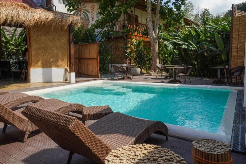 Outdoor pool area at El Nido Focus Rooms with wicker lounge chairs, small tables, and lush tropical greenery surrounding a modern building featuring thatched and wooden elements.